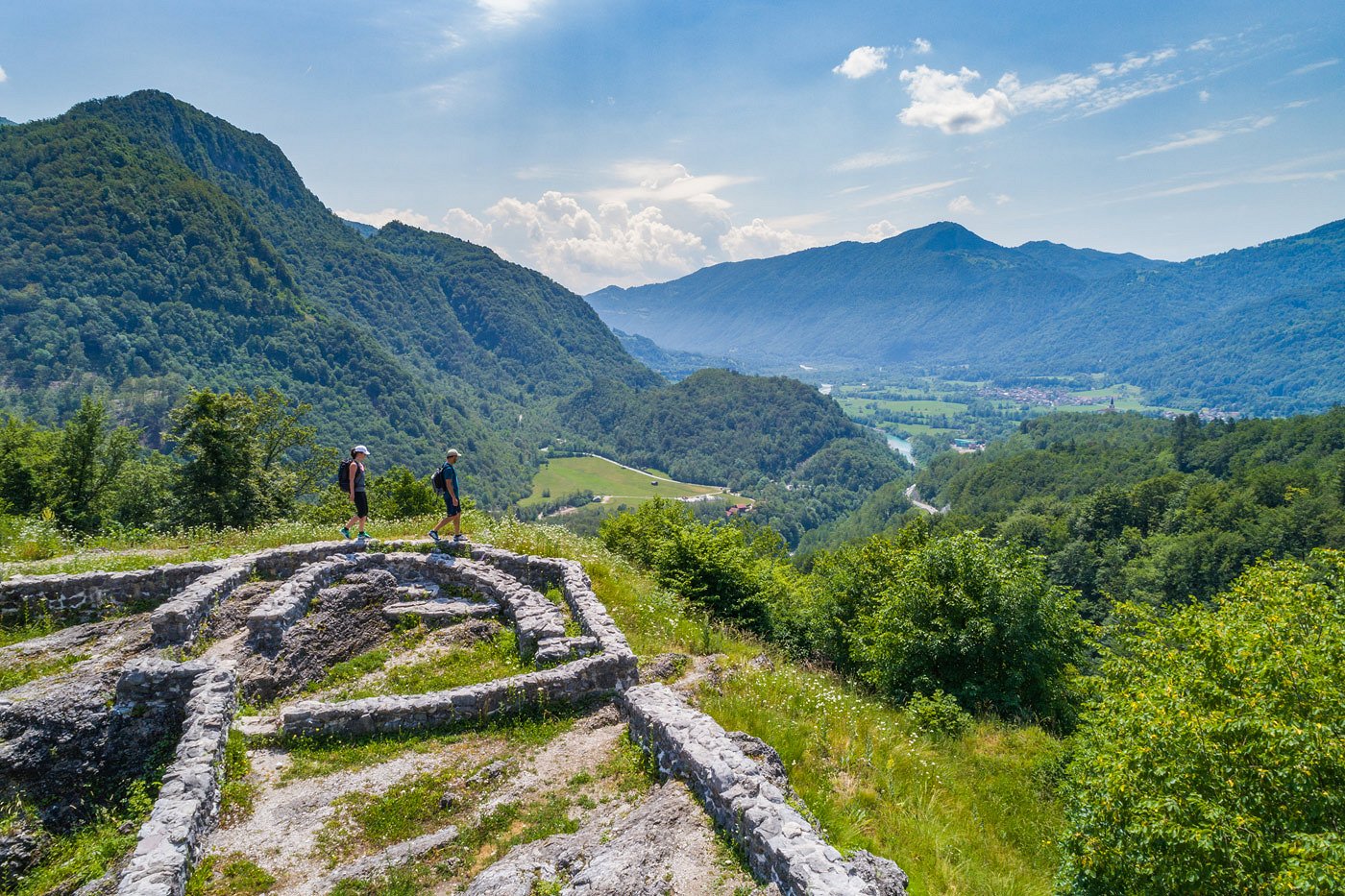 Hikers hike along the remains of the wall and enjoy the view of the valley in which the emerald green Soča River flows.