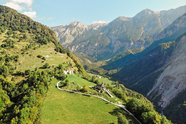 Sul lato sinistro dell'immagine, su un pendio erboso illuminato dal sole, si trovano la chiesa commemorativa dello Spirito Santo a Javorca e una fattoria vicina. Il prato è circondato da un bosco di alberi decidui ingialliti. A destra si apre la vista su una cresta montuosa soleggiata coperta di bosco, mentre nella parte inferiore si vede un pendio sabbioso in ombra, interessato da una frana.